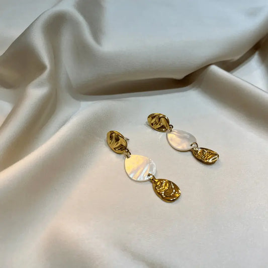 Boucles d'oreilles pendantes en acier inoxydable dorées avec détails en nacre, posées sur un fond en tissu