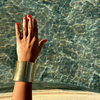 Main ornée d'un bracelet manchette en acier inoxydable au bord de l'eau, capturant la lumière naturelle pour une brillance dorée éclatante.