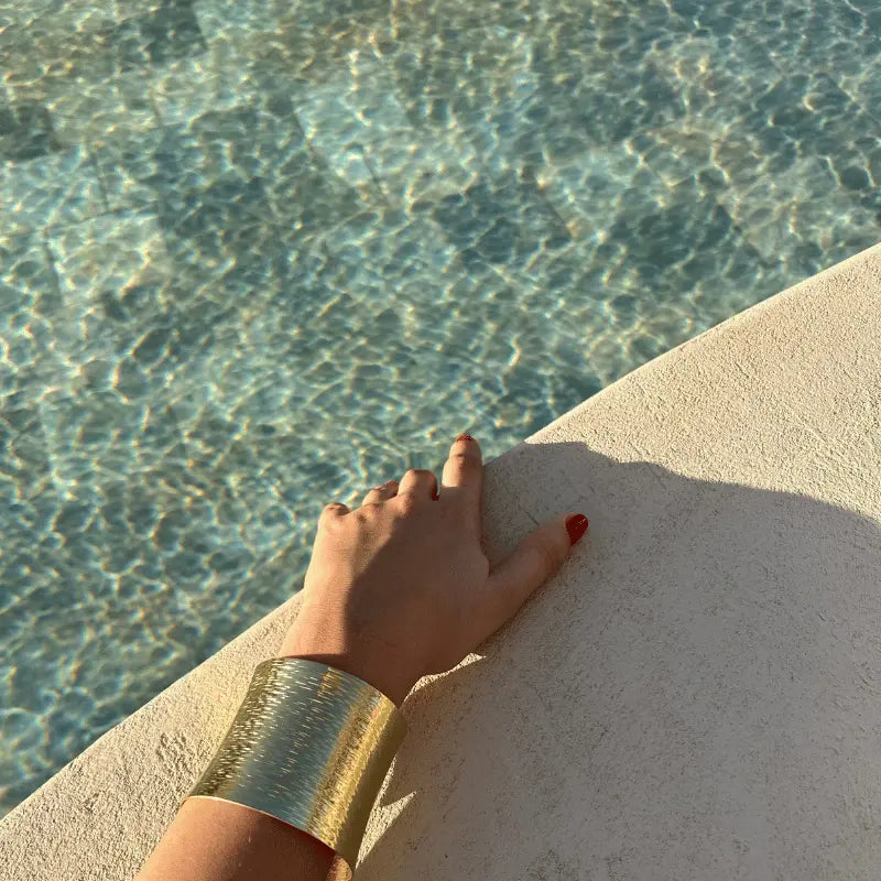 Bracelet manchette en acier inoxydable porté près de l'eau, mettant en valeur sa texture dorée brillante en plein soleil.