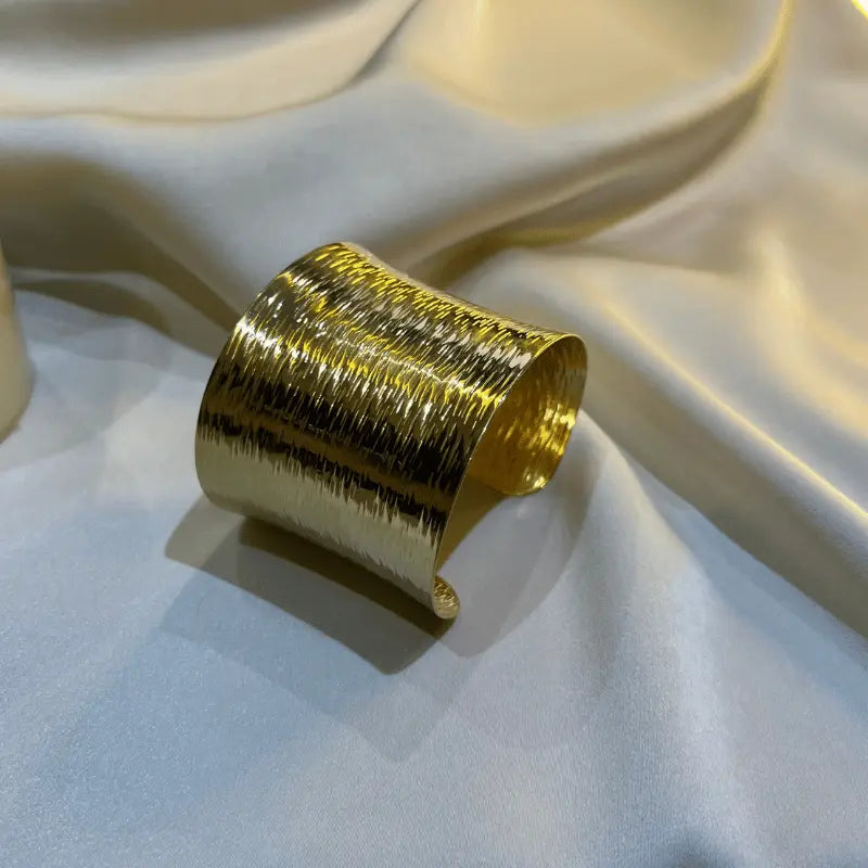 Bracelet manchette large en acier inoxydable, posé sur un tissu soyeux, reflétant une finition dorée élégante